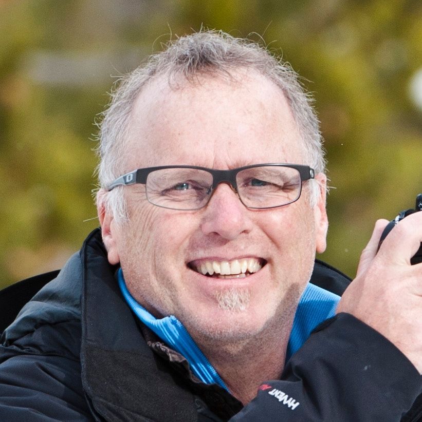 Man wearing glasses smiling while holding a camera outdoors.