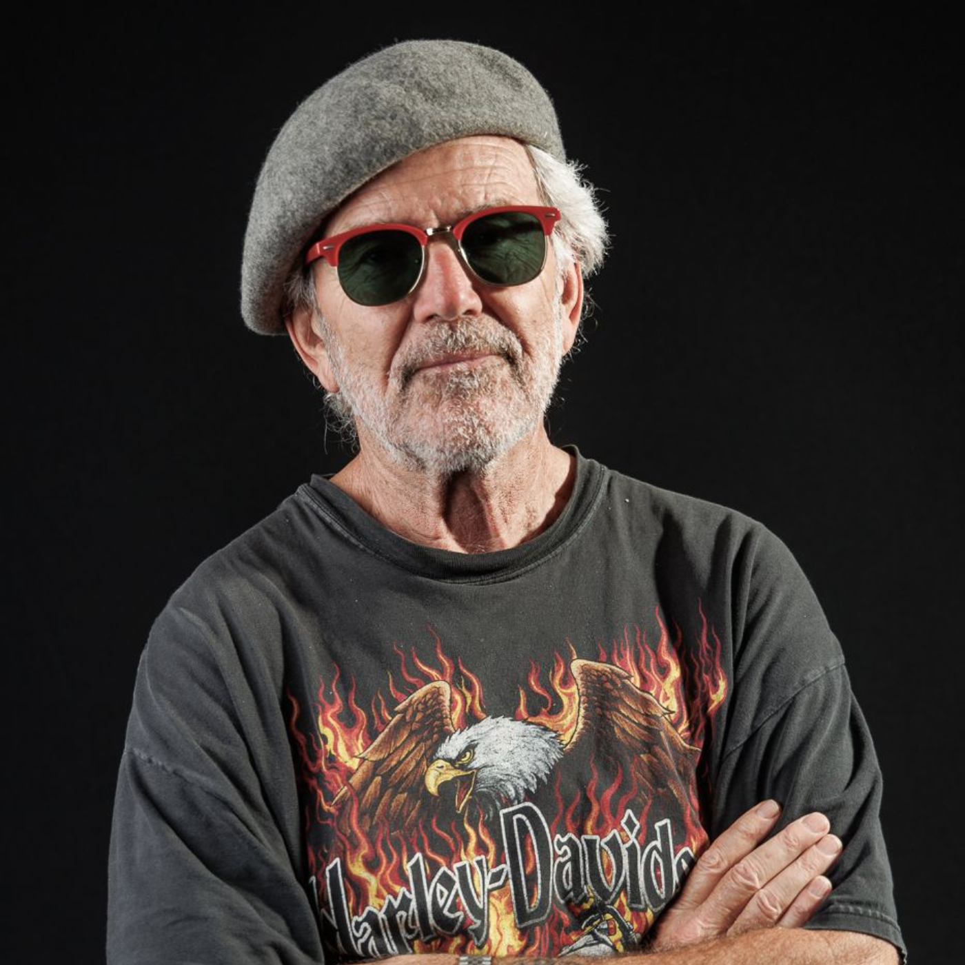 Man in sunglasses, beret, and Harley-Davidson t-shirt with arms crossed, in front of black background.
