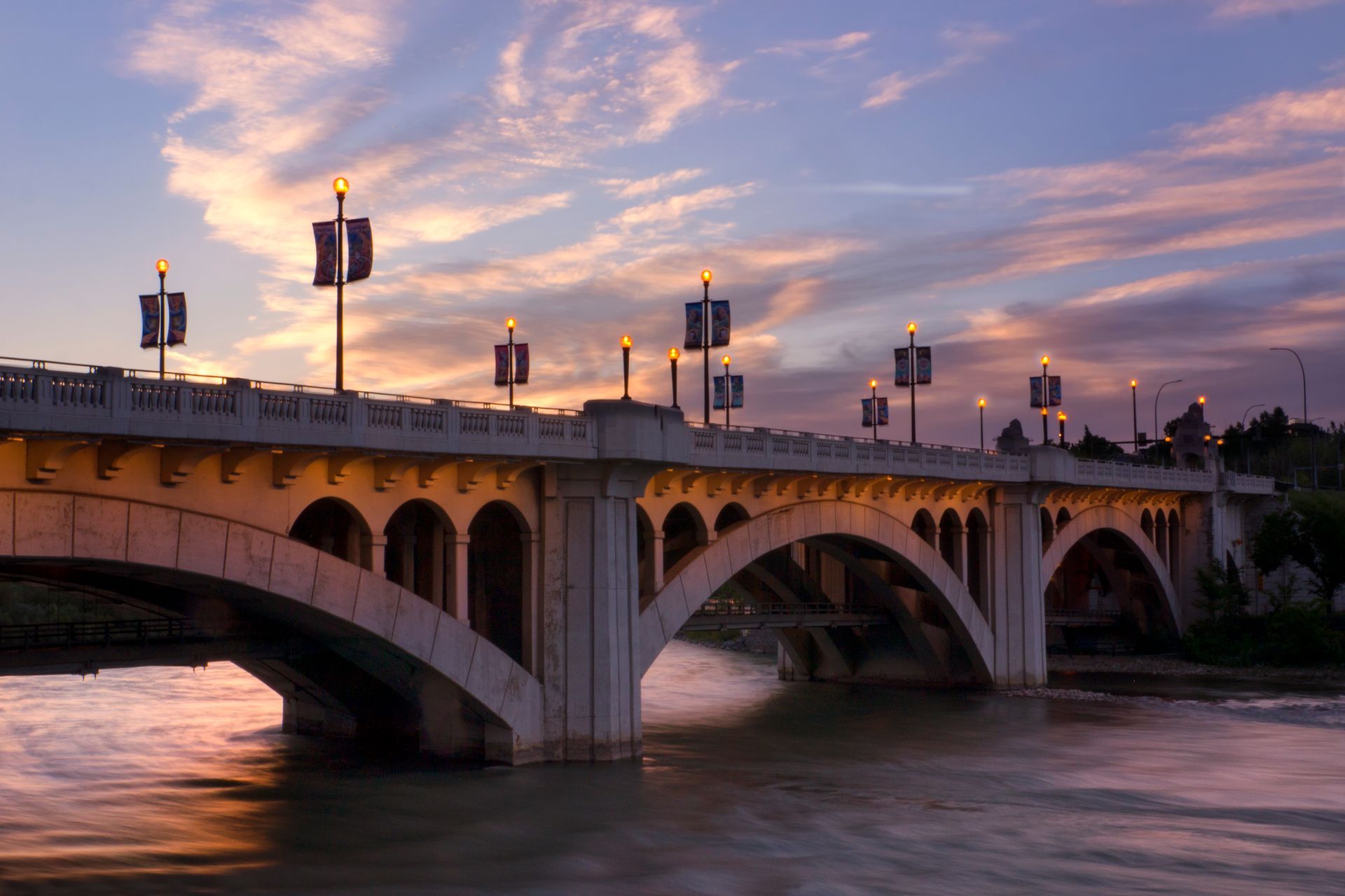 Stone bridge over river at dusk; banners line the structure; colorful sky.