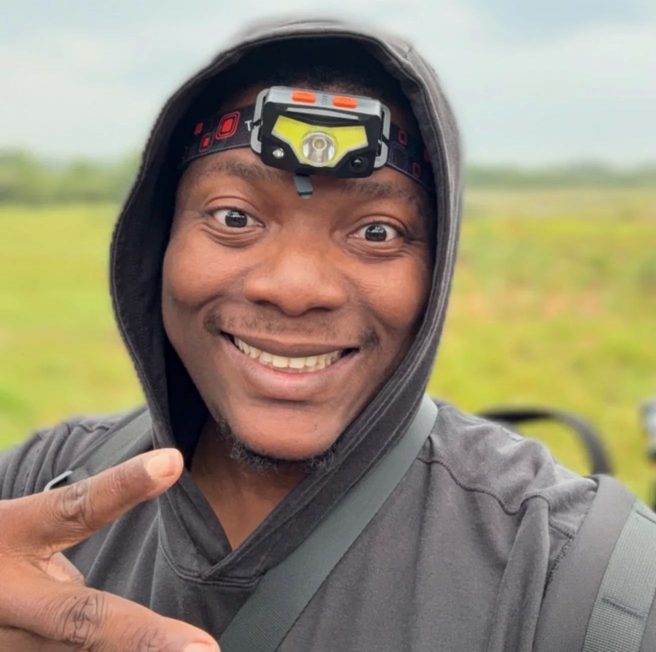 Man in hooded sweatshirt with headlamp, smiling and making peace sign outdoors.