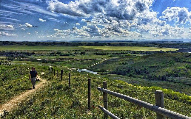 Person hiking on a grassy trail overlooking a valley. Cloudy sky. Green and brown landscape. Fence in foreground.