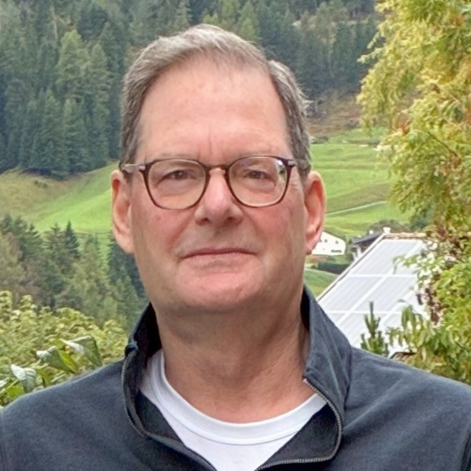 Man wearing glasses in front of a green mountain, wearing a black jacket and white shirt.