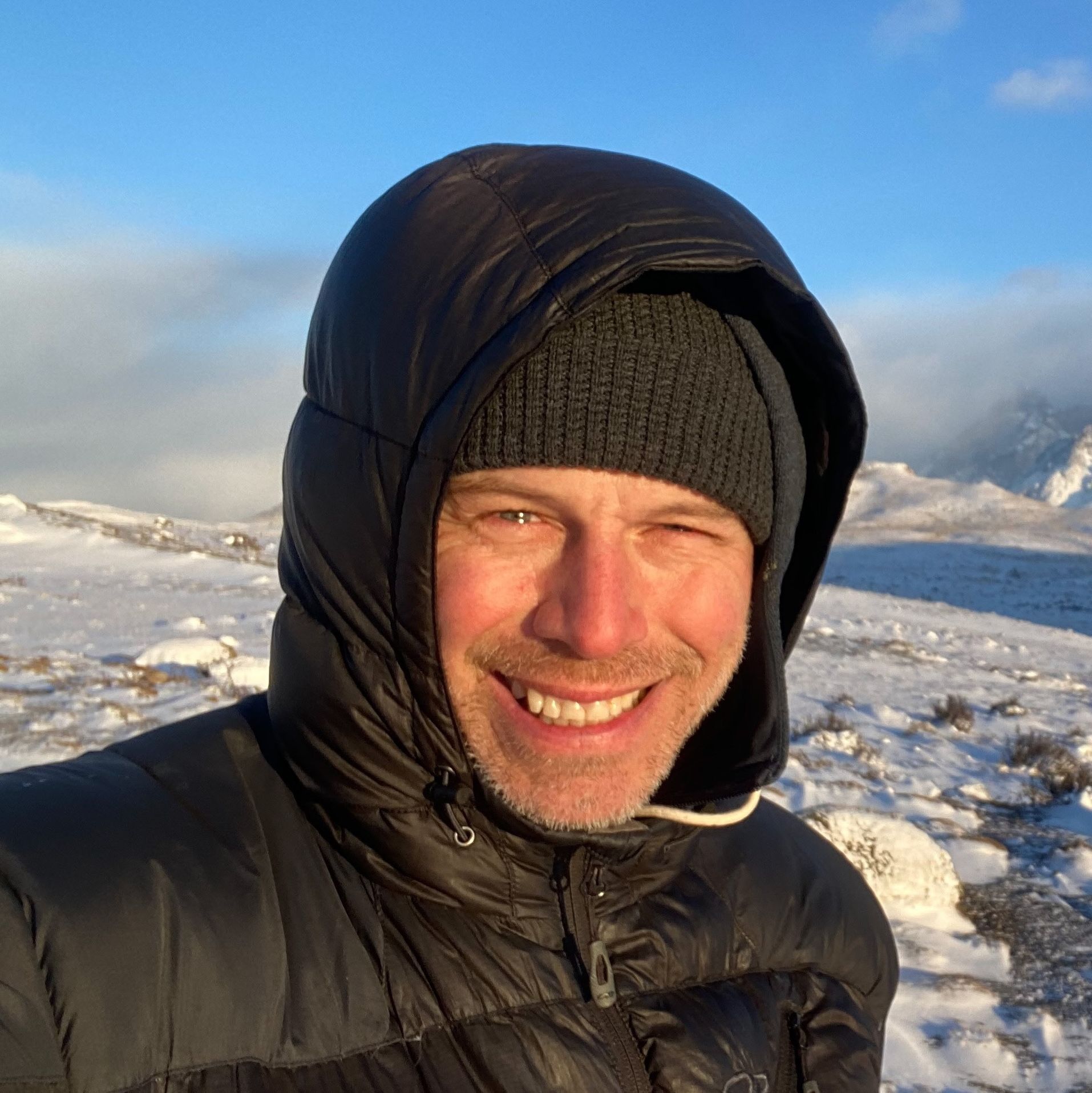Man smiles, bundled in winter gear, snowy mountain backdrop.