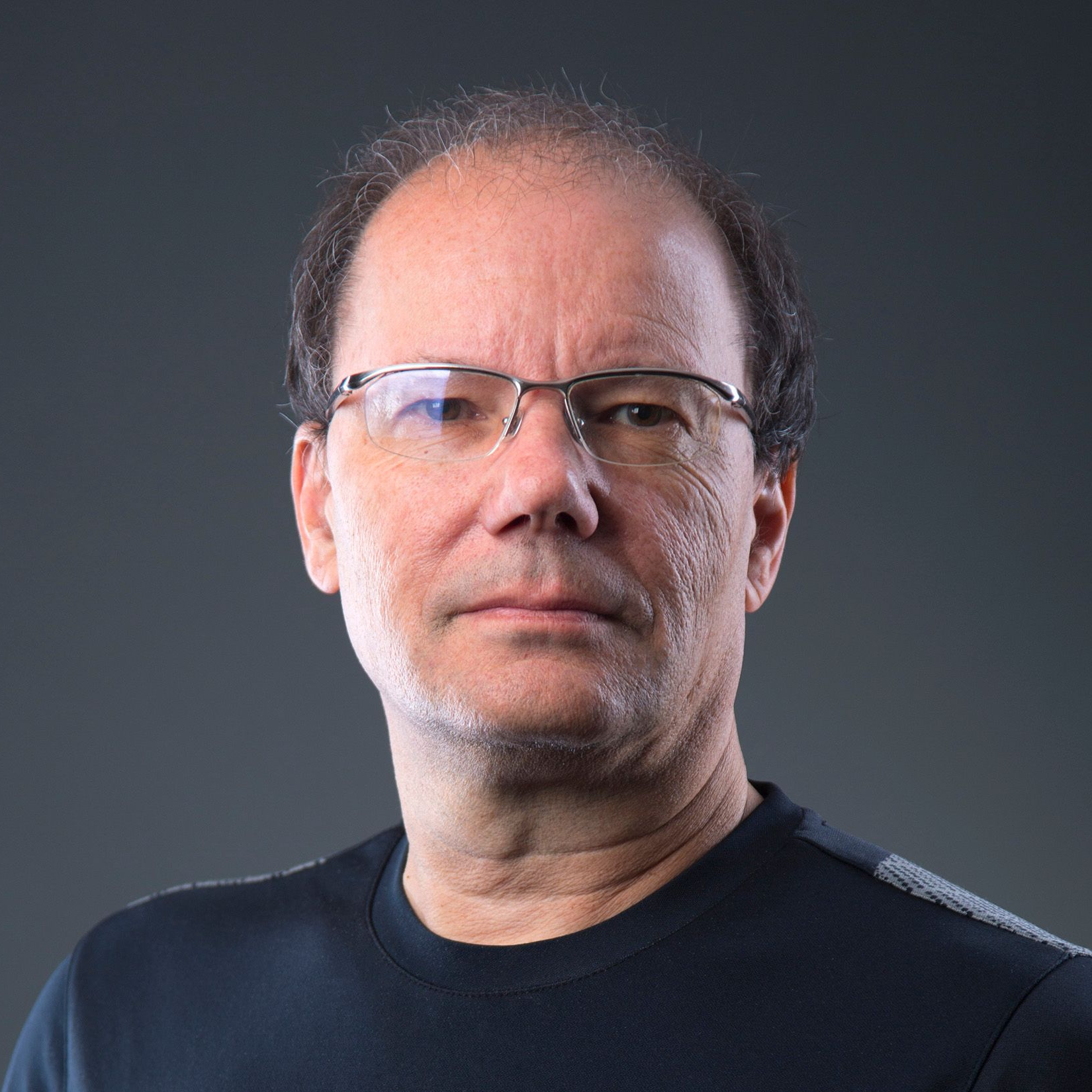 Man with glasses and a black shirt against a gray background, looking forward.