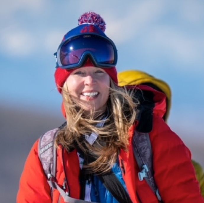 Woman in red jacket and hat with goggles smiles, outside in a snowy environment.