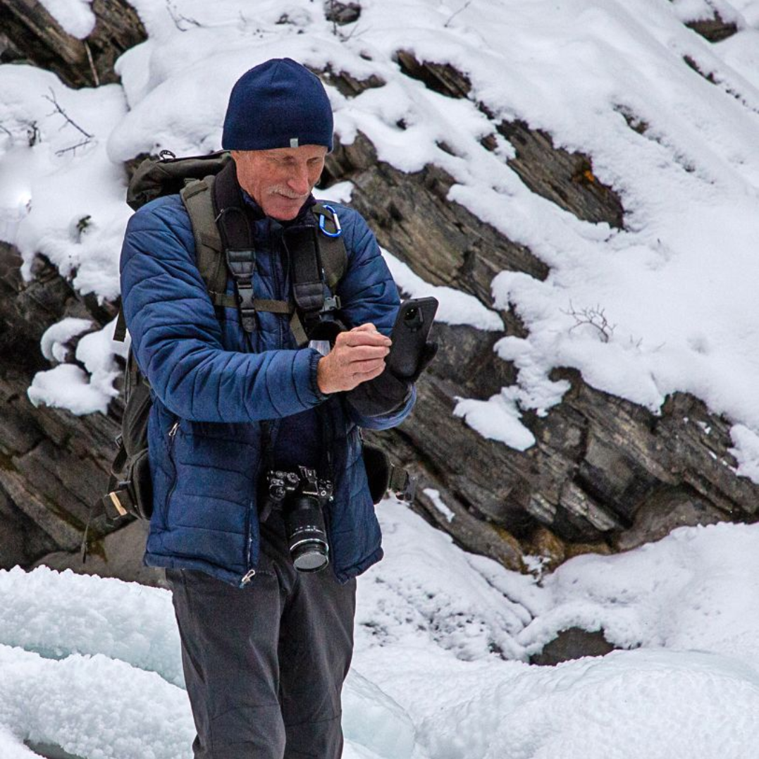Man in blue jacket adjusts gloves, camera around his neck, snowy rocky background.