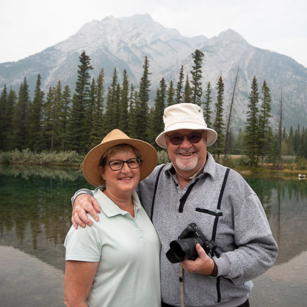 Couple poses outdoors with mountain and lake backdrop. Man holds camera; woman wears glasses and a straw hat.