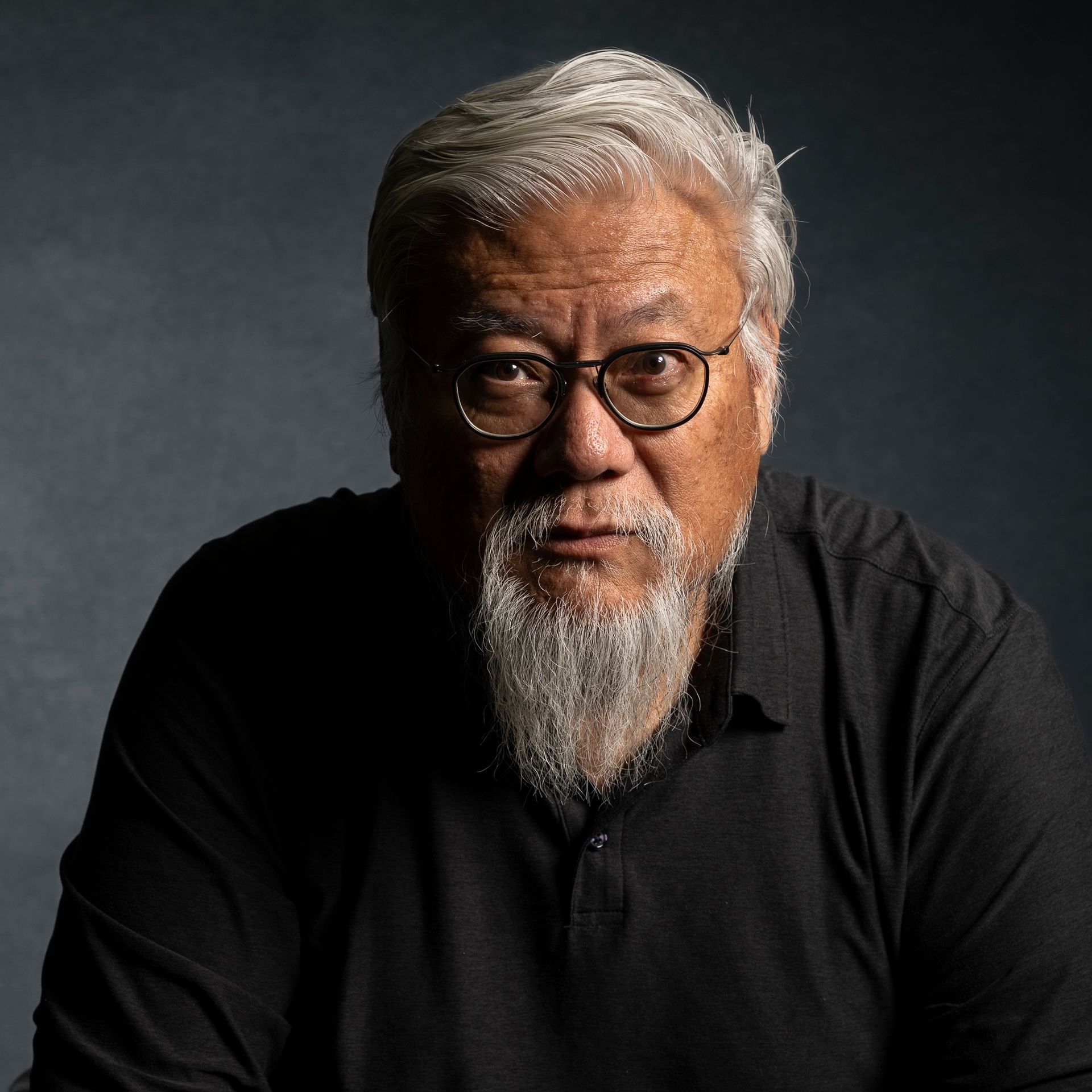 Man with gray beard and glasses, looking directly at the viewer against a dark background.