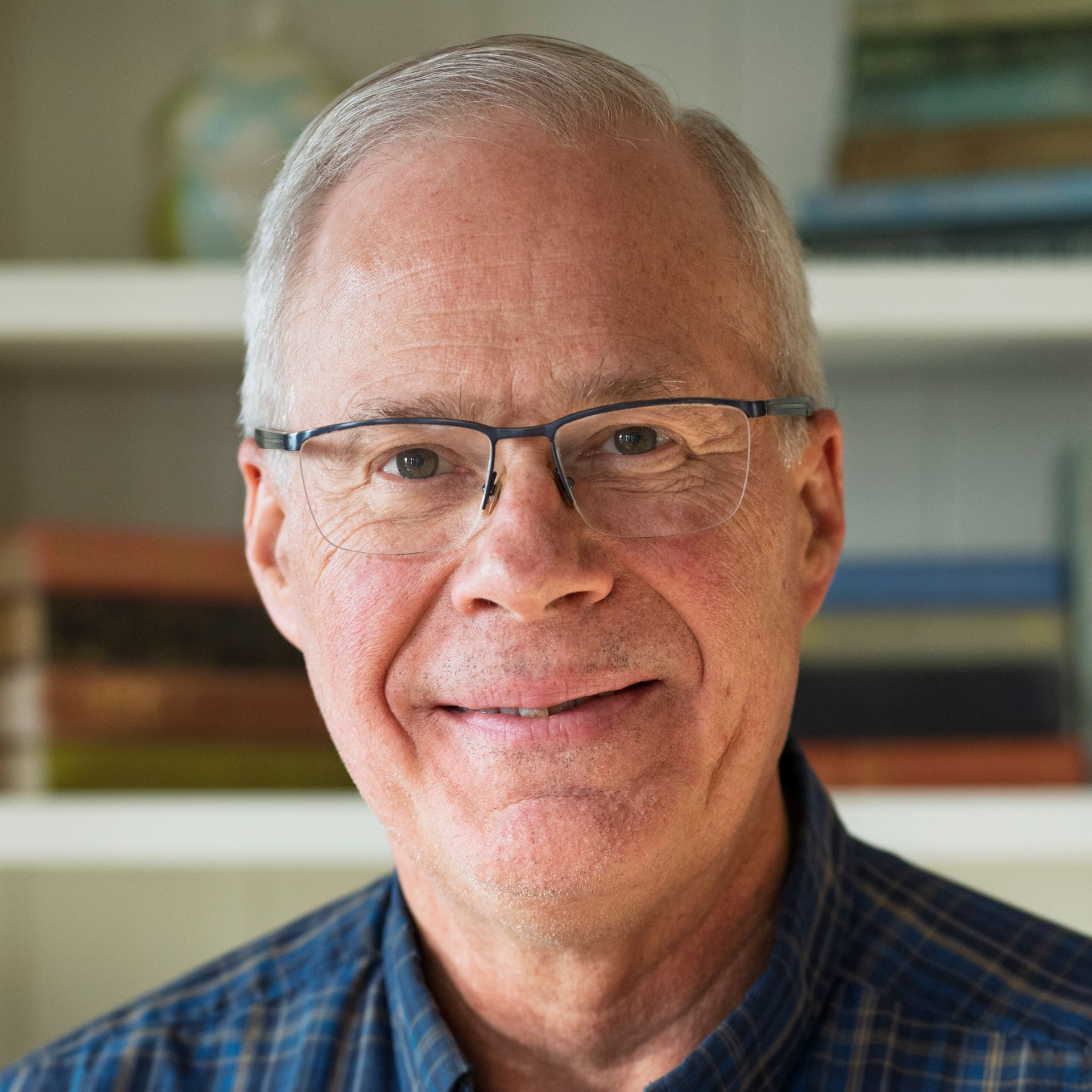 Man with glasses smiles, close-up. He wears a blue plaid shirt, set in front of a shelf of books.