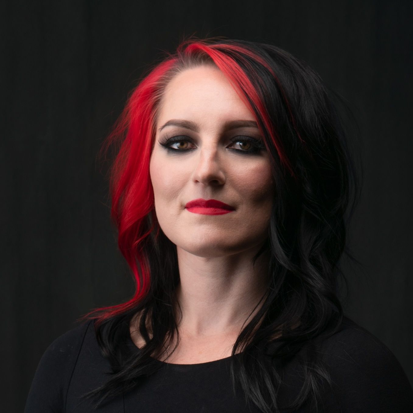 Woman with red-streaked black hair, dark makeup, and red lipstick, against a black background.
