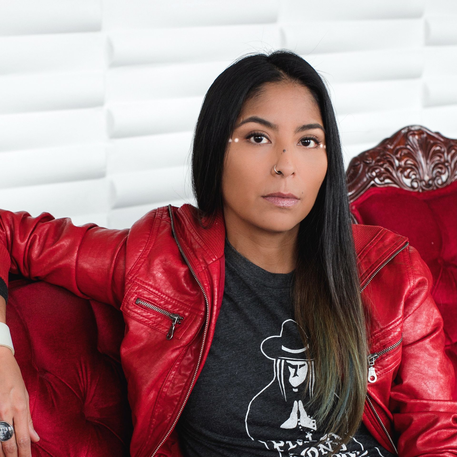 Woman in red jacket and graphic tee sits on a red velvet chair, looking at the camera.