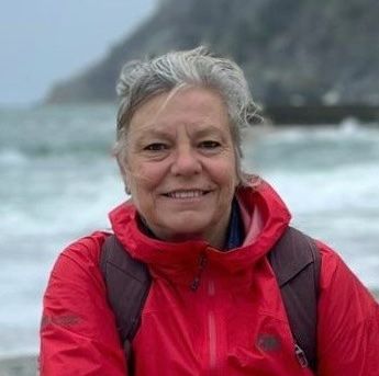 Woman in a red jacket smiles, standing near a body of water with a mountain in the background.
