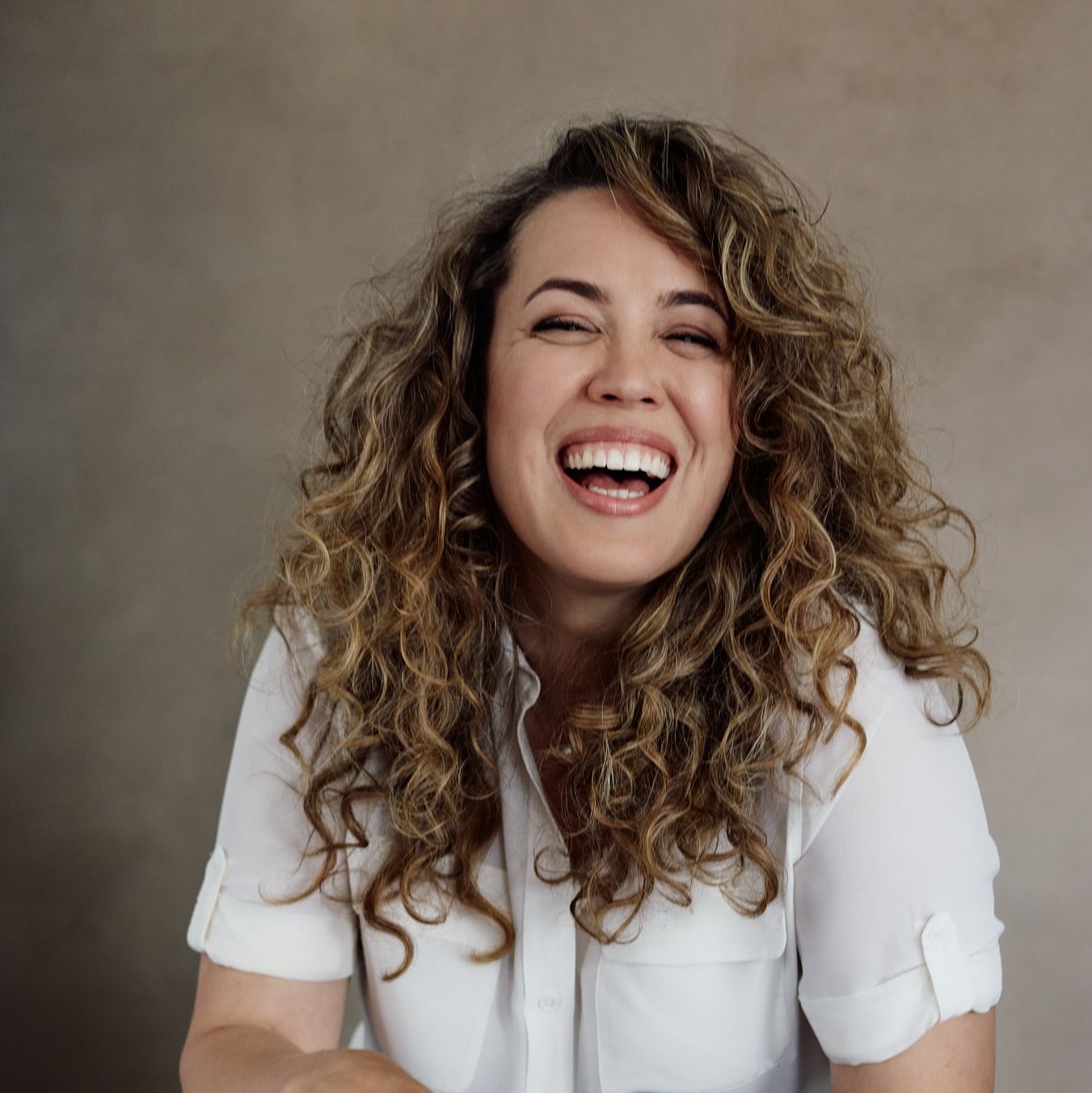 Woman with curly hair laughing, wearing a white shirt, against a neutral background.