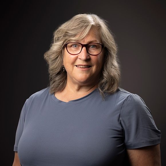 Woman with gray wavy hair and glasses smiles. Wearing a blue shirt, against a dark background.