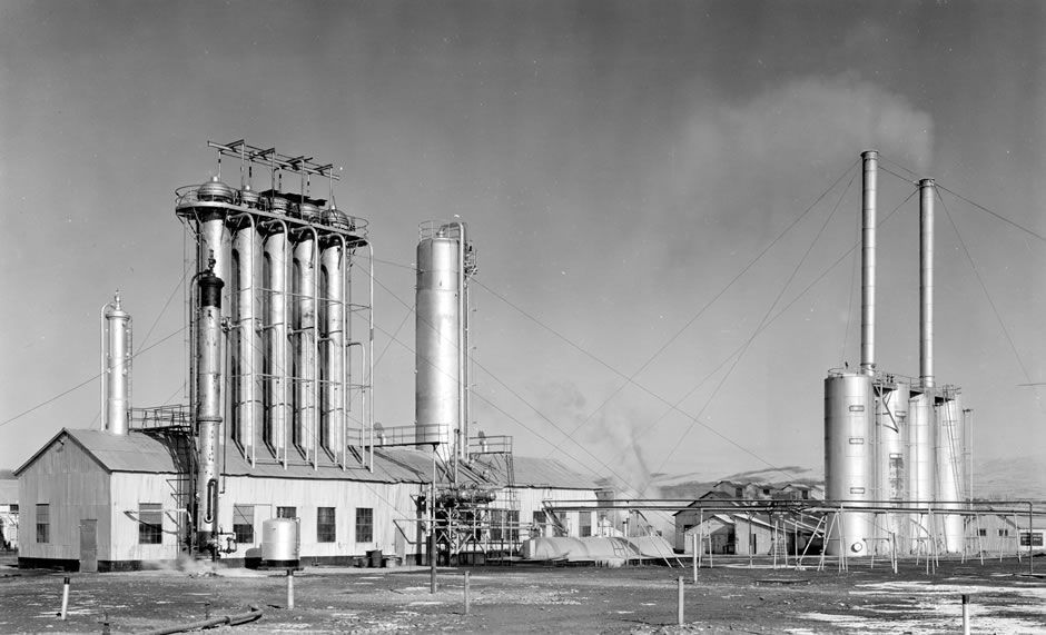 A historic industrial plant with tall cylindrical structures, pipes, and smokestacks against a cloudy sky.
