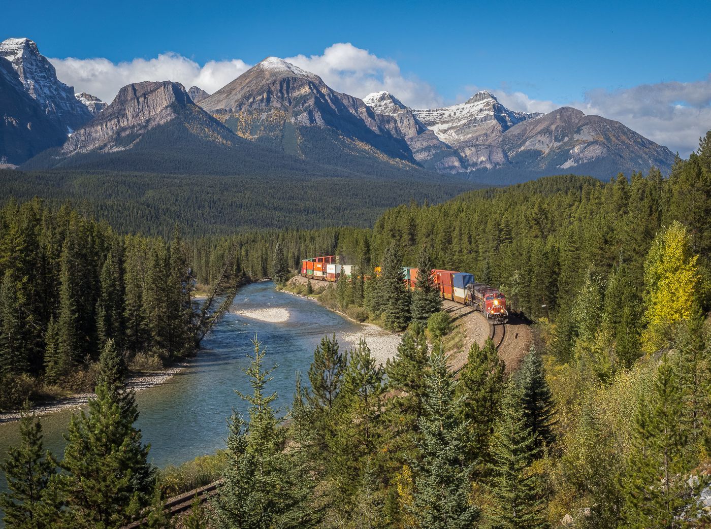 Train with cargo containers travels alongside a river in a mountain landscape.