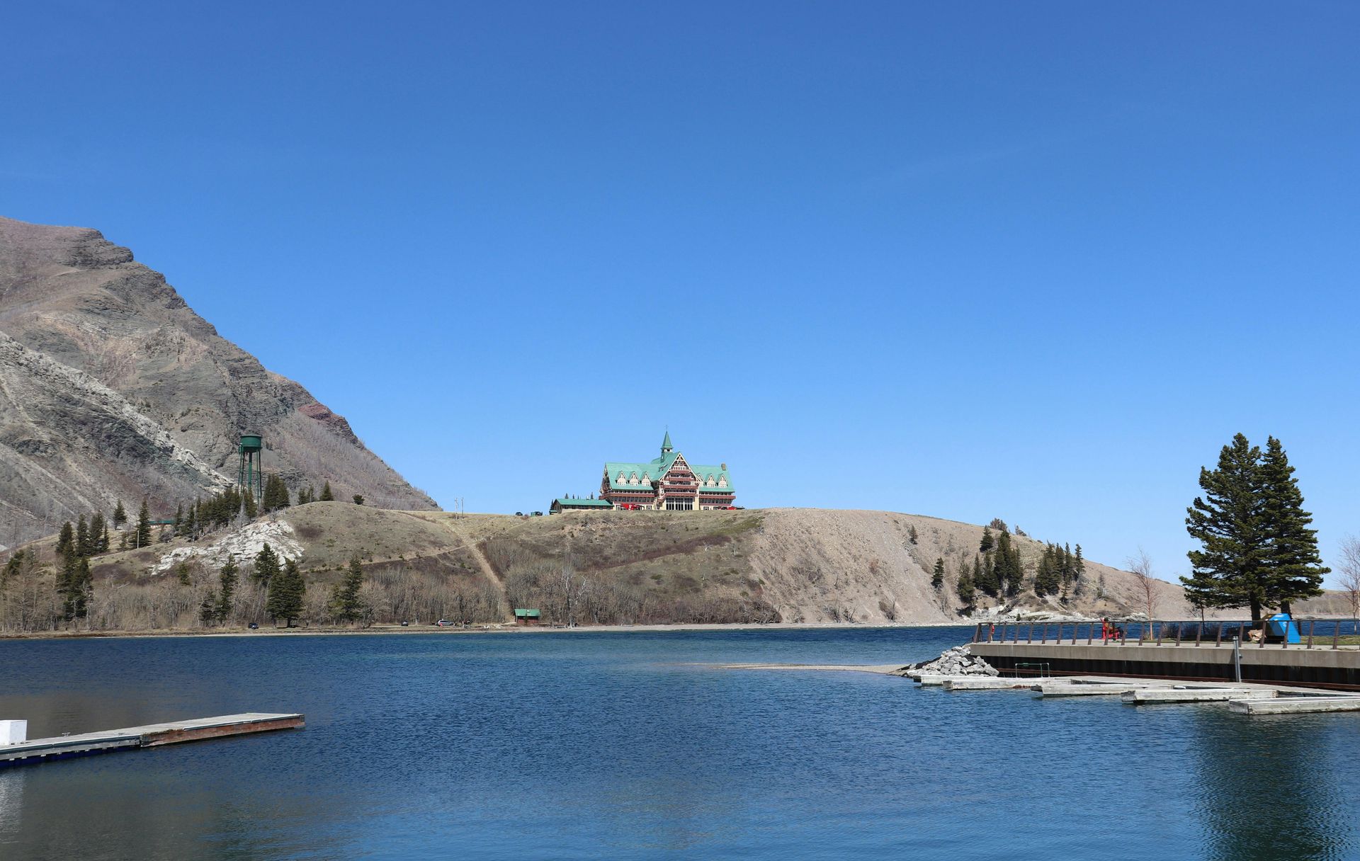 Blue lake with a brick building on a hillside under a bright blue sky. A mountain and dock are visible.