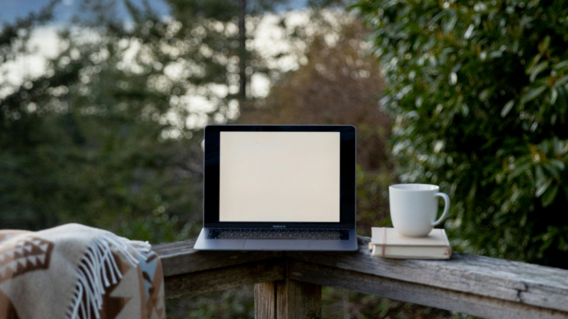 Laptop on a wooden railing with a coffee cup and books, overlooking a blurred green forest.
