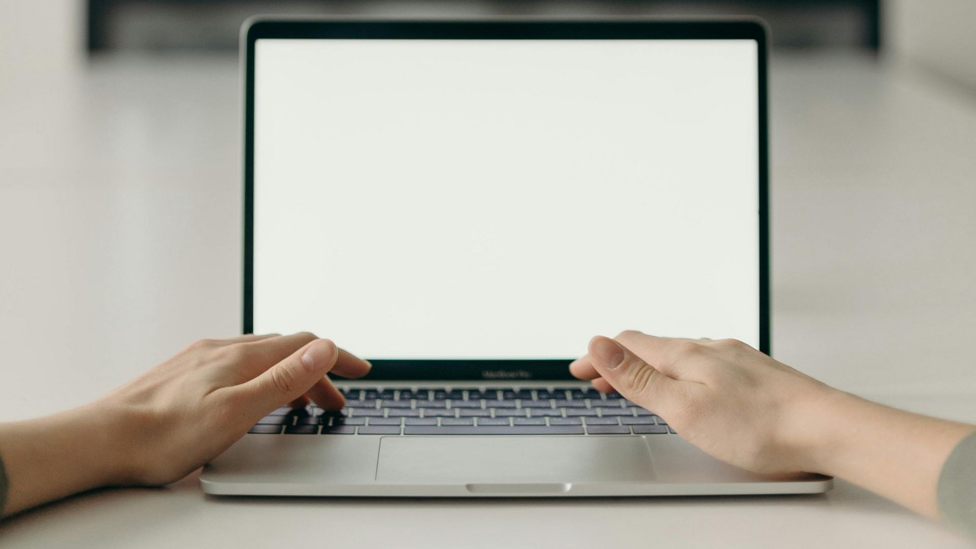 Hands typing on a silver laptop with a blank white screen, on a white surface.