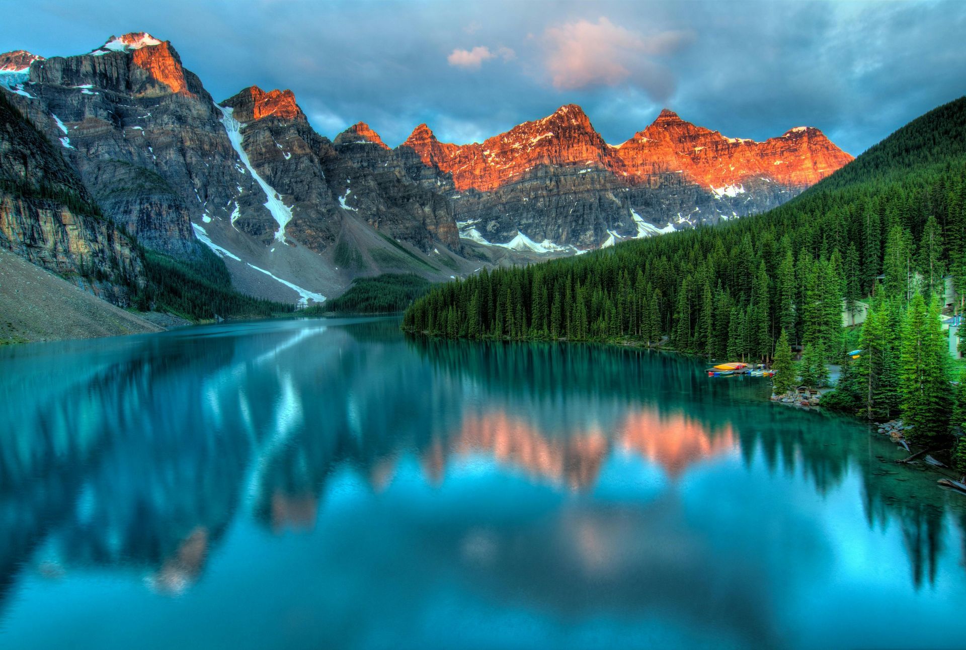 Emerald lake reflecting snow-capped mountains at sunrise. Pine forest on the right.