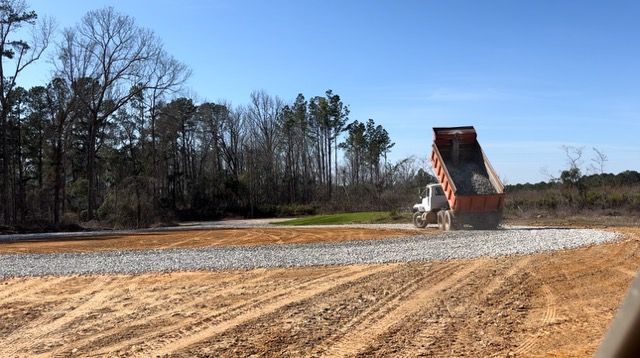 A dump truck unloading gravel on a dirt road, with trees and blue sky in the background.