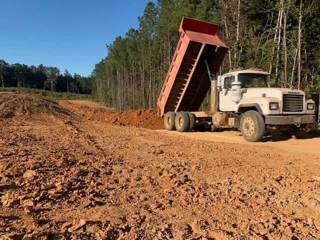 Dump truck unloading dirt onto an earthen road in a wooded area.