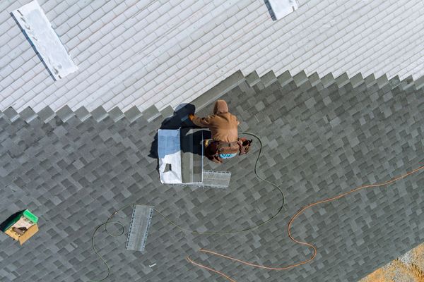 Roofer installing shingles on a roof. They are kneeling, working on a gray shingled roof, with tools and materials nearby.