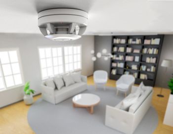 Smoke detector mounted on a white ceiling, overlooking a modern living room with light furniture and bookshelves.
