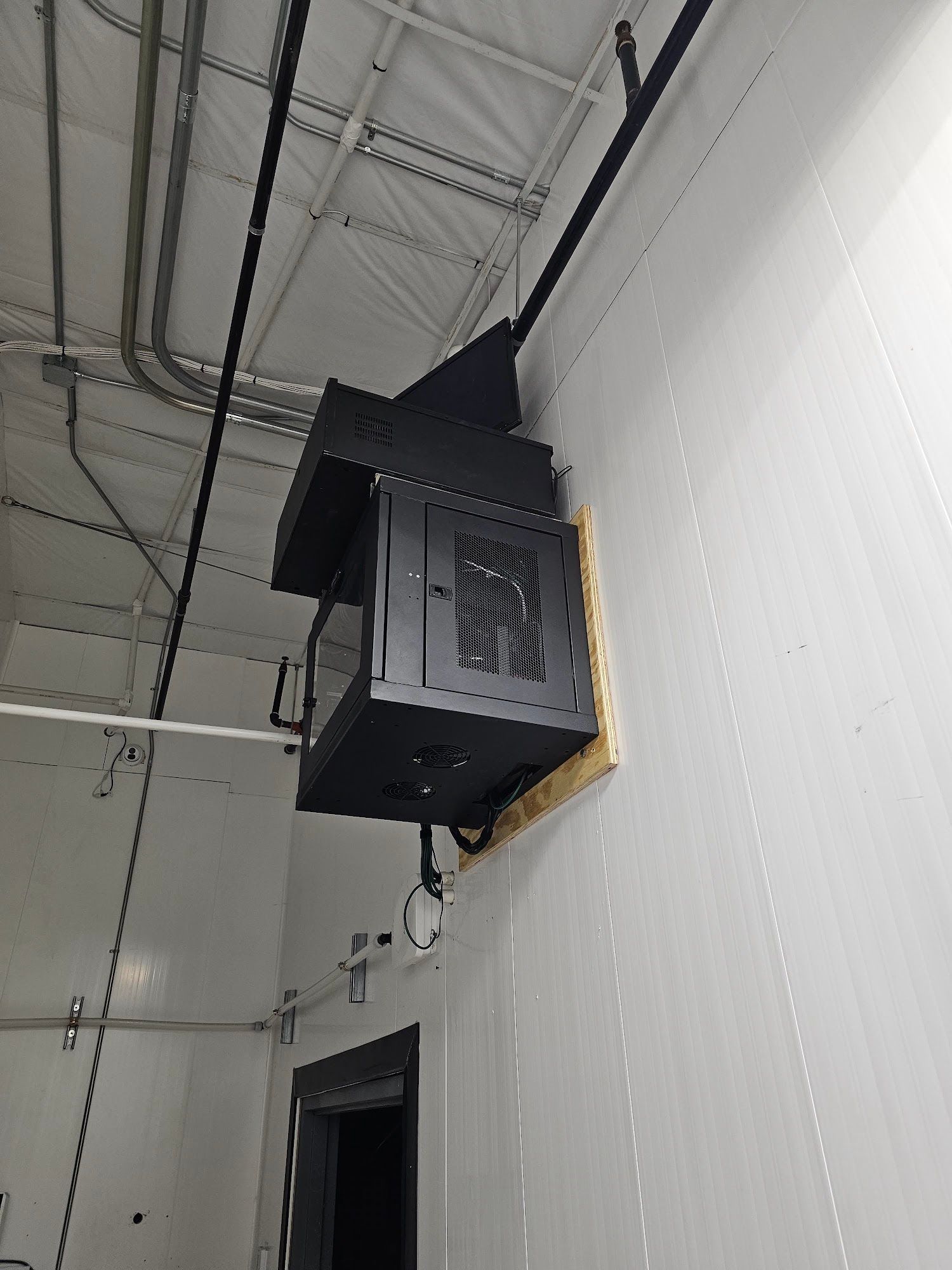 Black metal HVAC unit mounted on a white wall in a room, with electrical conduit visible.