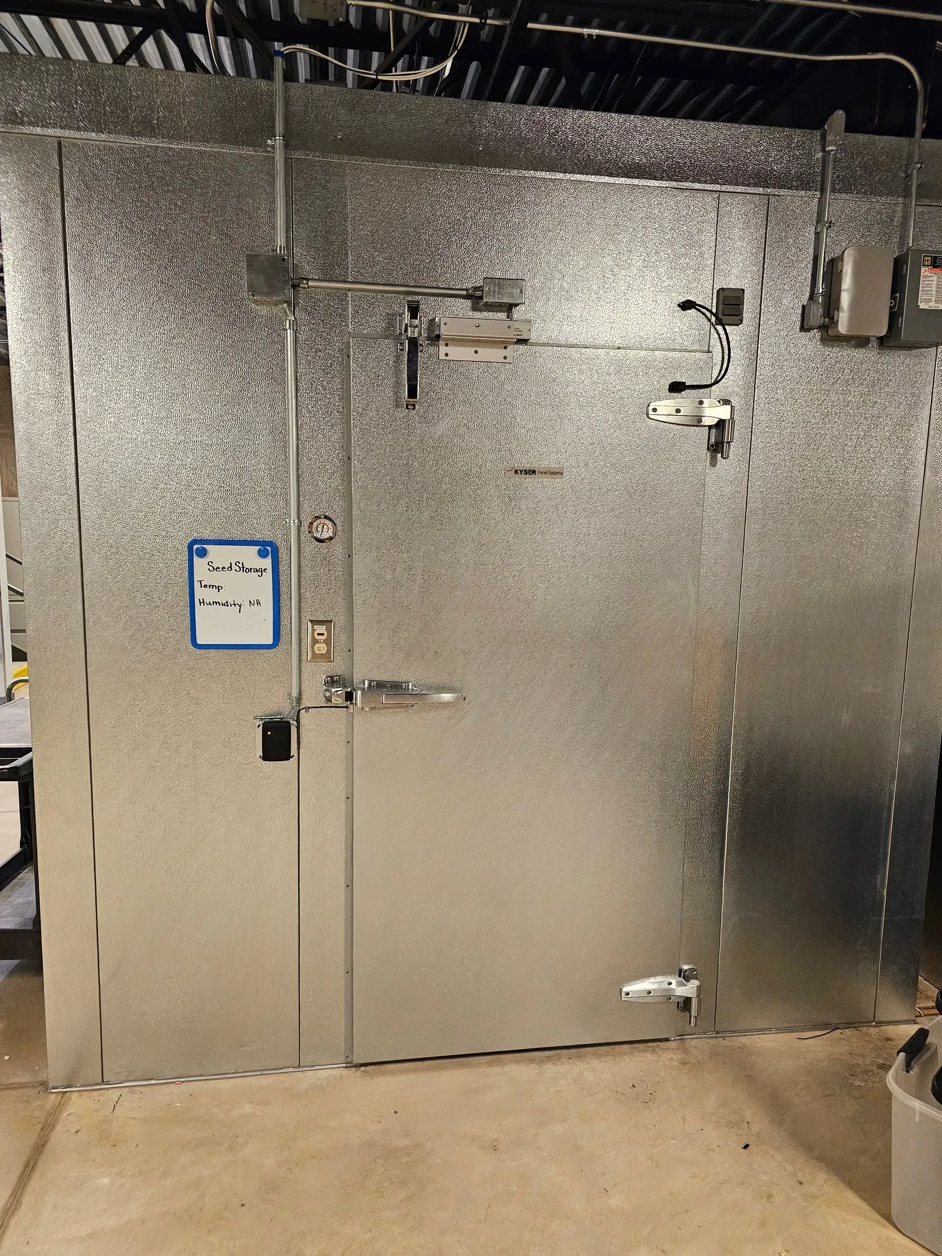 Metal walk-in refrigerator/freezer in a commercial setting; door has handles and latch; control box on the wall.