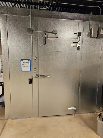 Metal walk-in refrigerator/freezer in a commercial setting; door has handles and latch; control box on the wall.