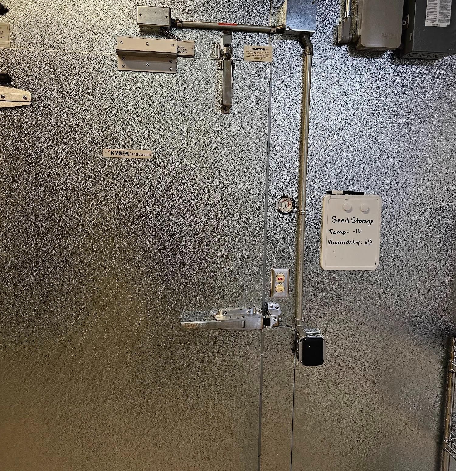 Silver-paneled walk-in cooler door with door hardware, electrical conduits, and a handwritten note.
