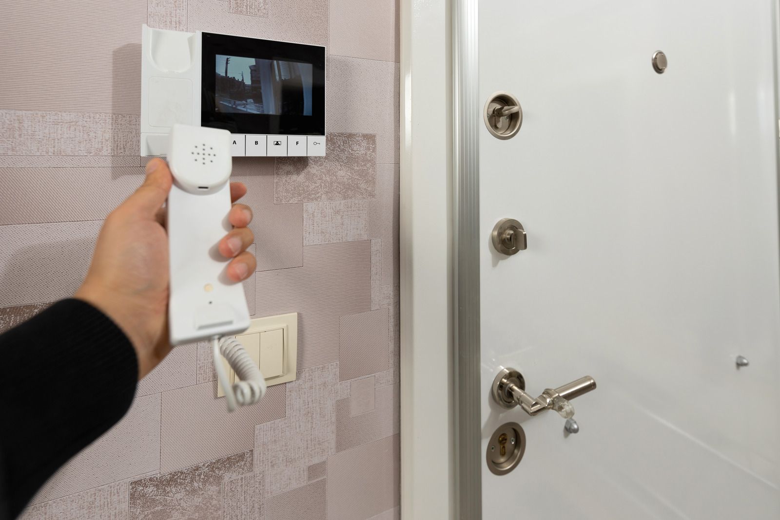 Person holding a white intercom handset near a door with a video screen.