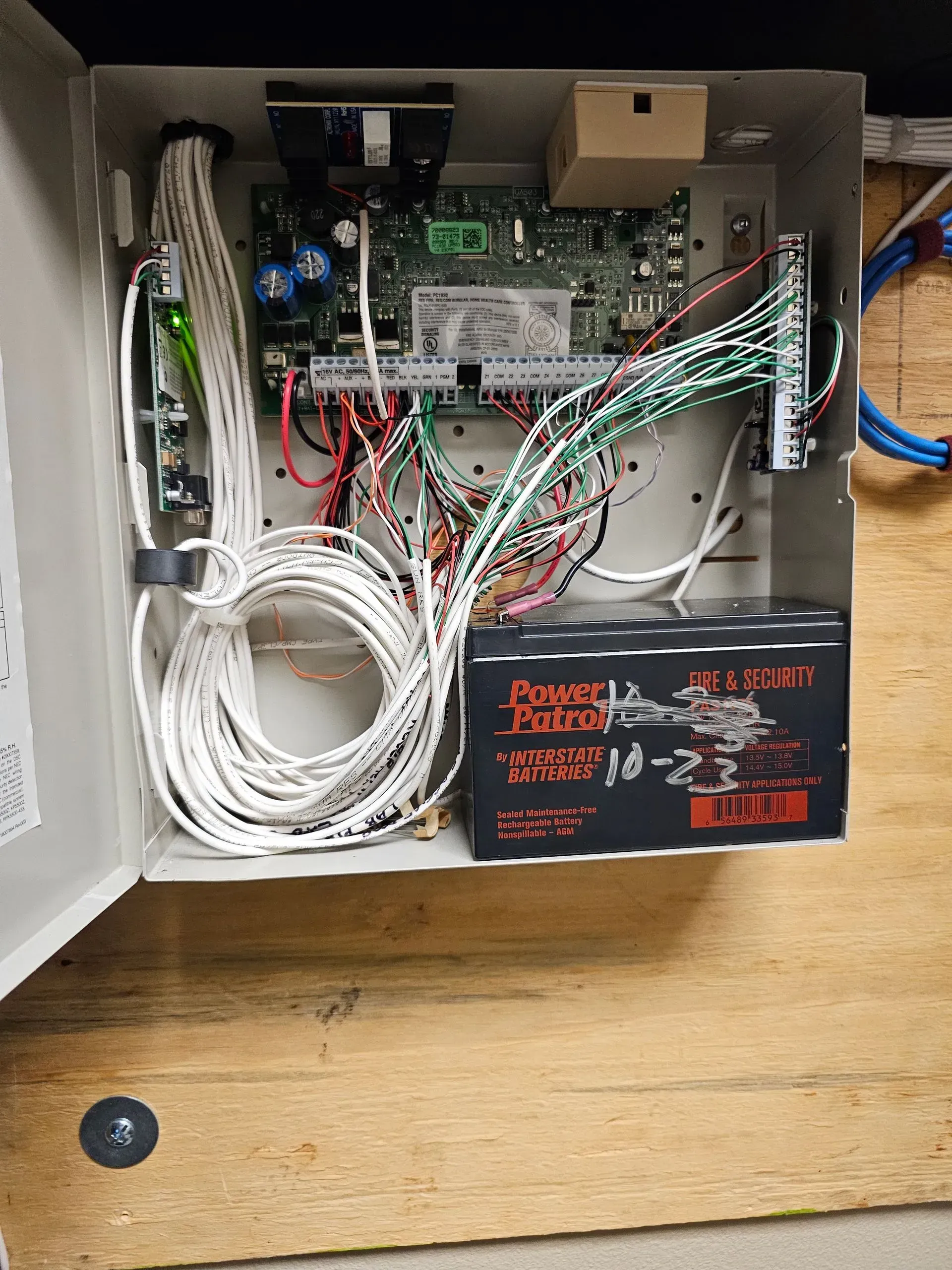 Open security system panel with wires, circuit board, and battery on a wooden surface.