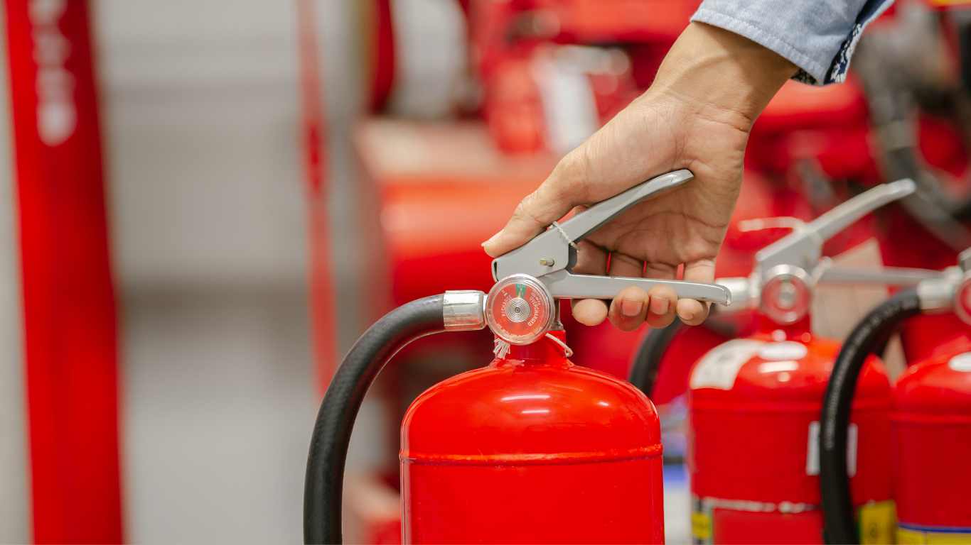 Person's hand on the lever of a red fire extinguisher, with others in the background.