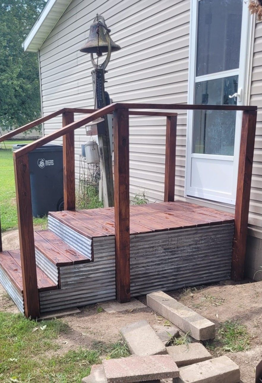 Wooden porch with steps, handrails, and corrugated metal siding. Red-brown wood. Exterior view.
