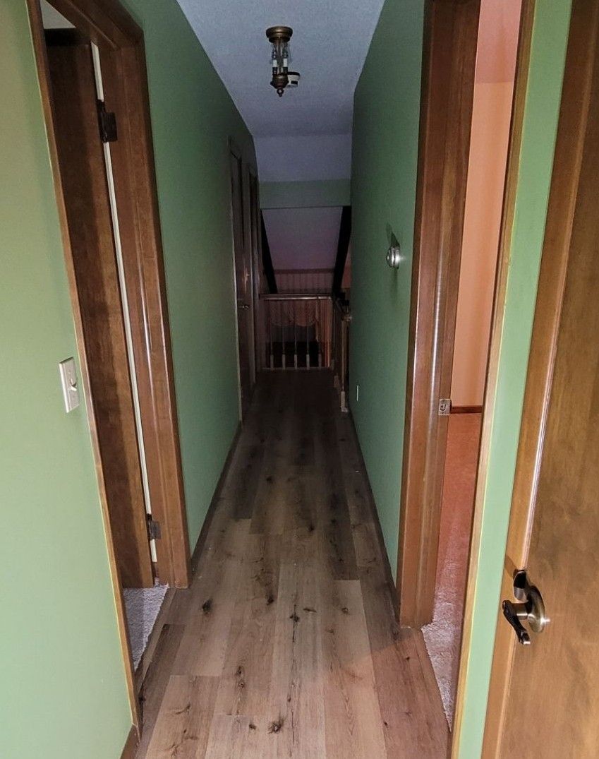 Narrow hallway with wooden floor, green walls, and doors on both sides, leading to a staircase.