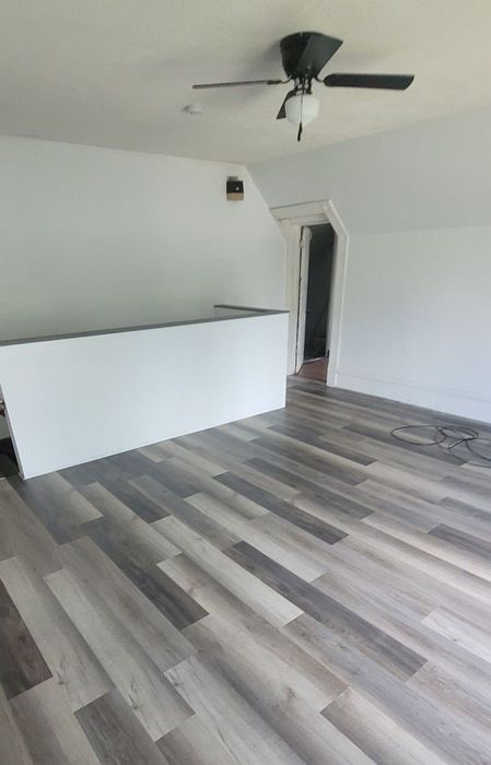 Room with gray wood-look flooring, white walls, a half-wall, and a ceiling fan. A doorway is visible.