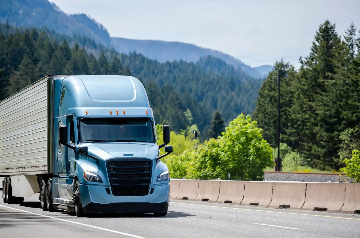 A blue semi truck is driving down a highway with mountains in the background.
