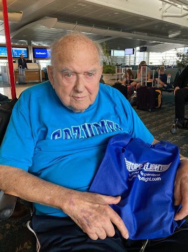 An elderly man is sitting in a wheelchair at an airport holding a blue bag.