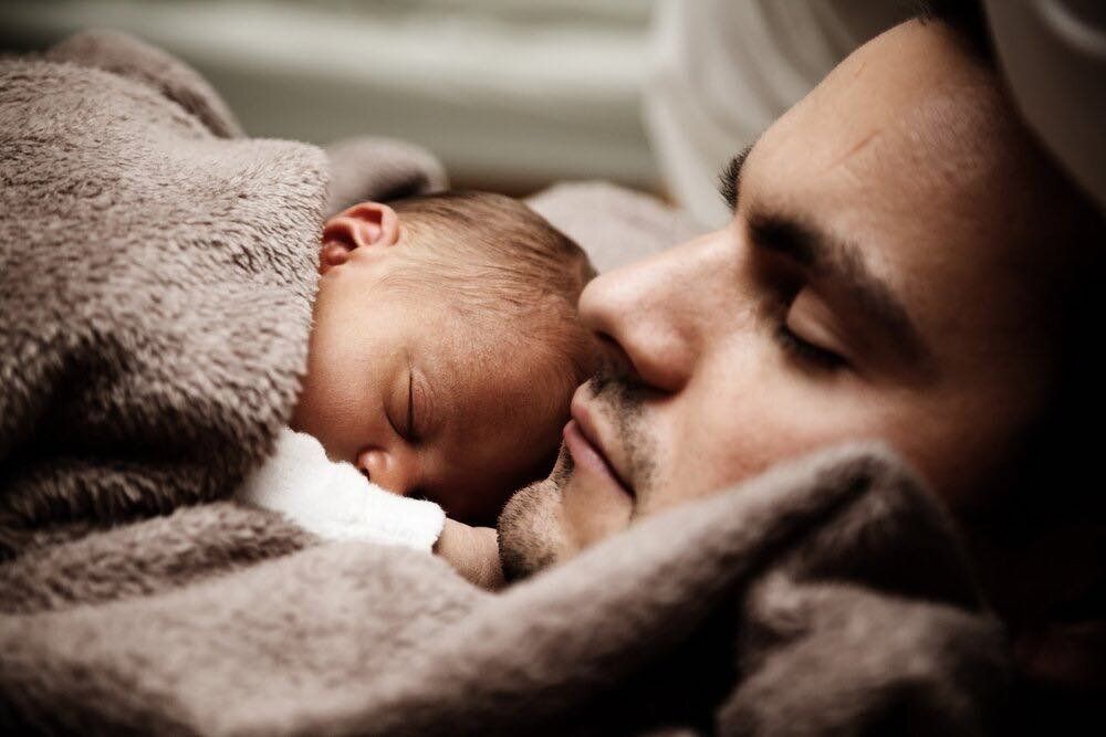 A man is sleeping with a baby wrapped in a blanket.