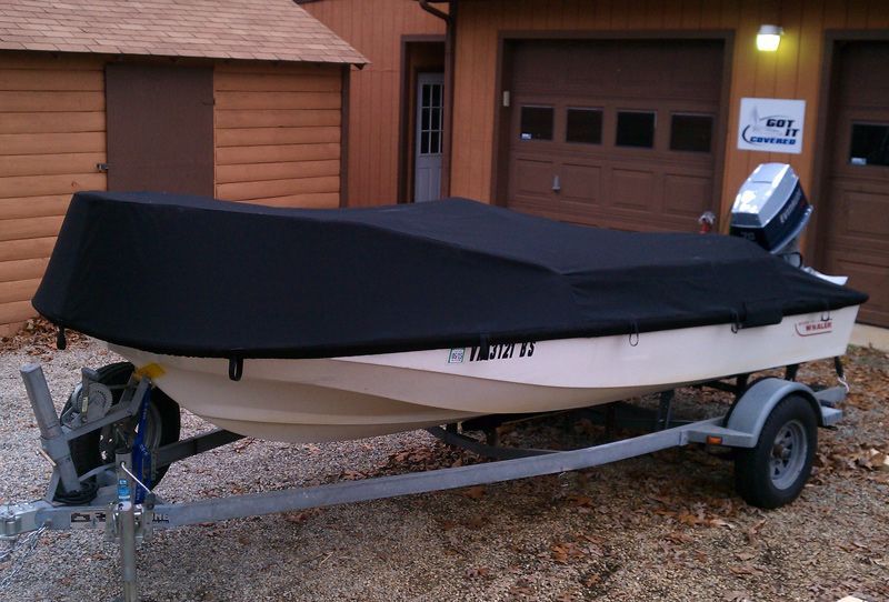 A small boat with a black cover on a trailer in front of a shed and garage.
