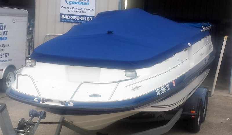 Boat on a trailer, covered with a blue tarp. White hull, black tires, parked in front of a building.