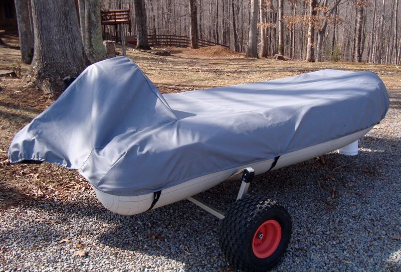 Covered boat on a trailer, gray cover, tan boat, black straps, red wheels, outdoor setting, trees in background.