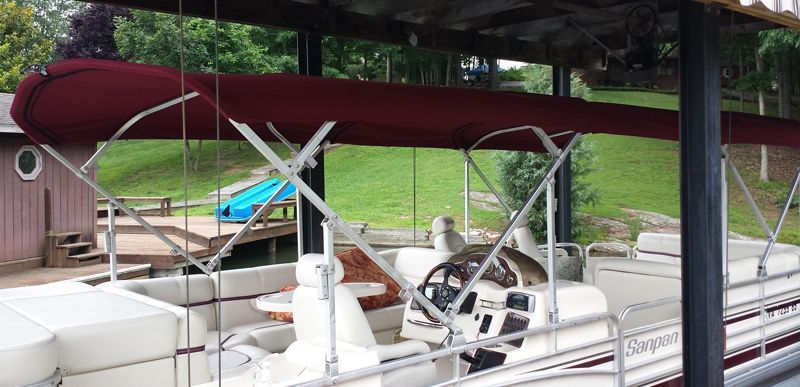 A maroon-topped pontoon boat under a boat dock with a steering wheel and white seating. Green grass in the background.