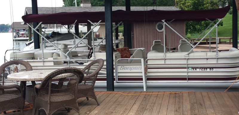 A pontoon boat docked under a covered area, with a table and chairs on a wooden deck.