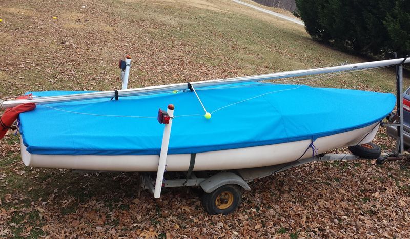 Small sailboat on trailer, covered with a blue tarp. Two white poles support the tarp, green grass in the background.