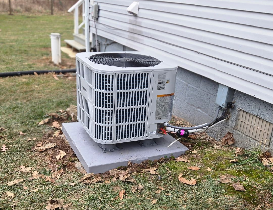 Two men inspecting an outdoor heating unit on a house. One points while the other holds papers.