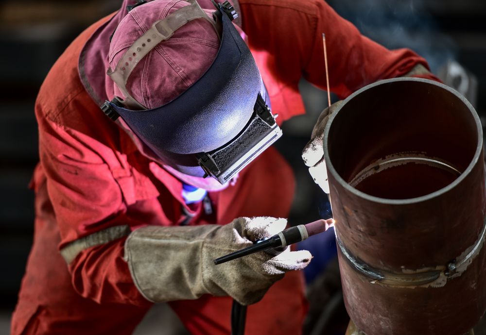 Welder in Red Coveralls Welding a Metal Pipe, Wearing a Helmet and Gloves — EB Metalworks in Somersby, NSW