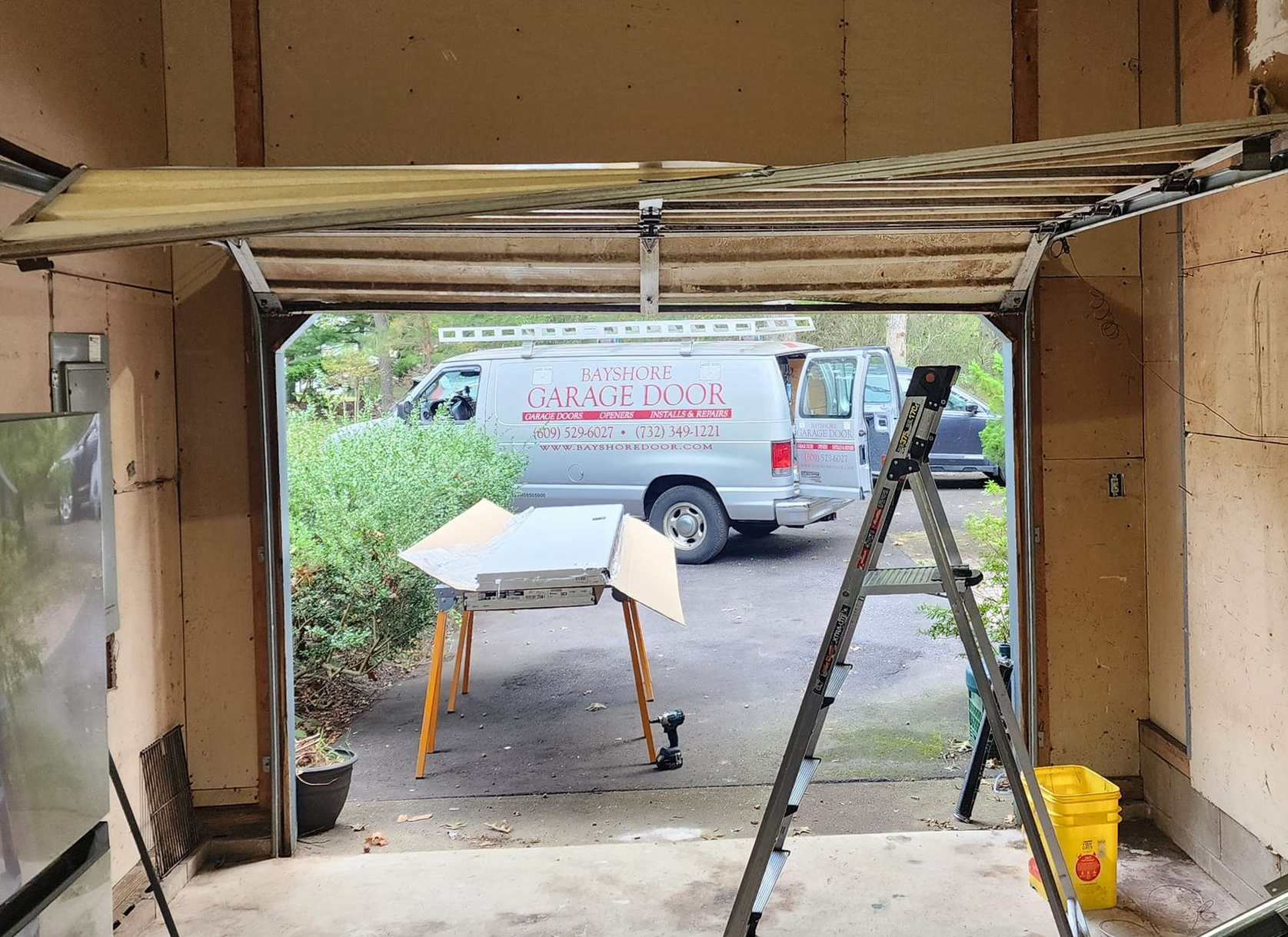 A detached garage door hangs in its tracks with a service van and tools visible in the driveway outside.